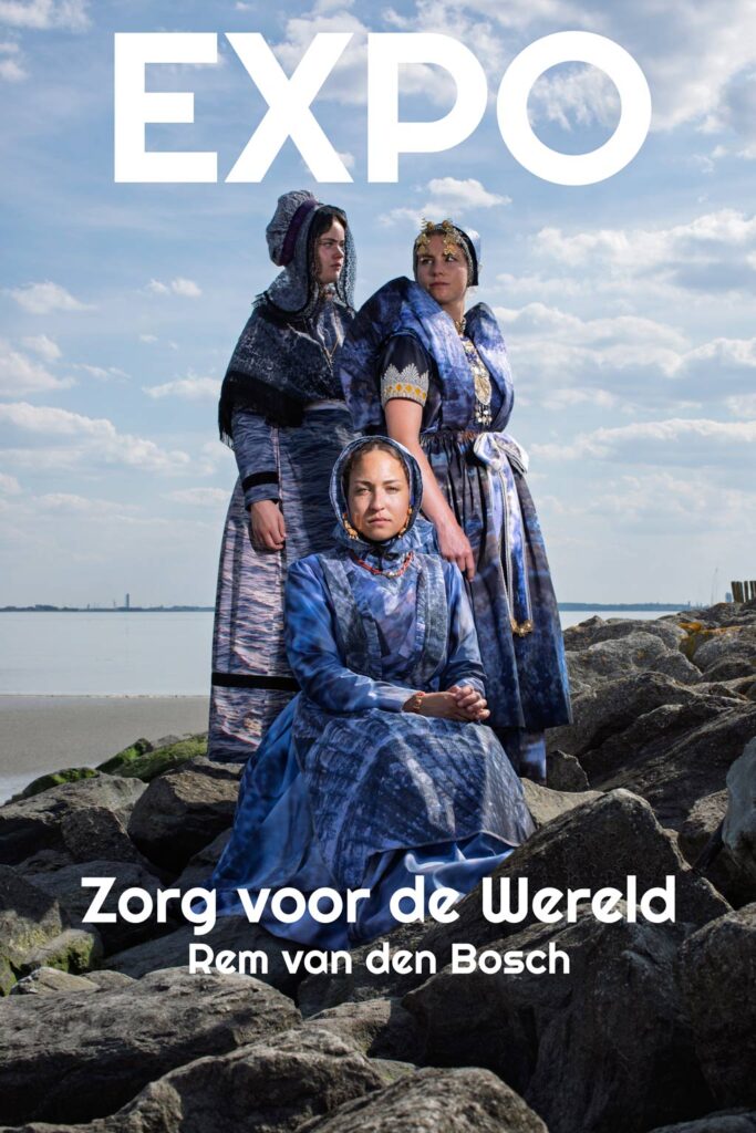 Drie vrouwen in Zeeuws-Vlaamse klederdracht poseren op een rotsachtige kust voor de expositie 'Zorg voor de Wereld' van Rem van den Bosch. De kostuums zijn gemaakt van duurzaam materiaal in diverse blauwtinten.